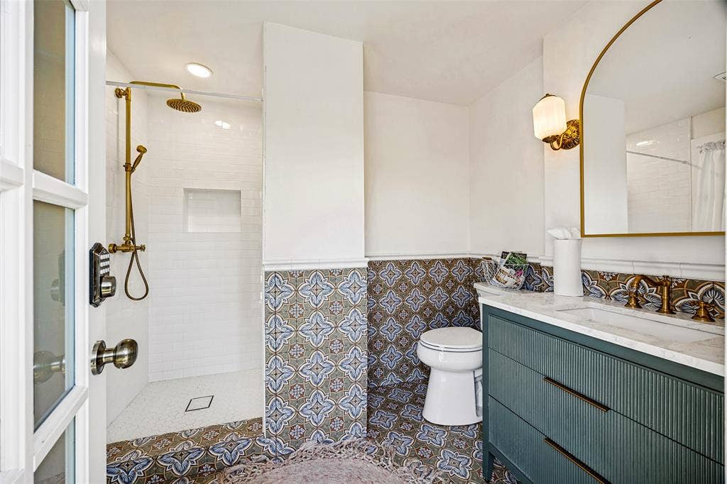 320 E 5th Street, Dallas, TX 75203 Walk-in shower with brass fixtures and recessed niche, patterned floor and wall tile, vanity with integrated sink and storage, arched mirror with sconce lighting