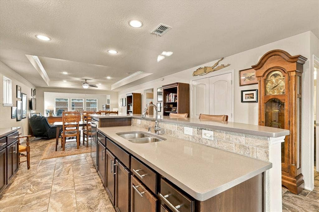 Kitchen featuring dark-finish cabinetry, neutral-toned solid surface countertops, a dual-basin stainless steel undermount sink, stone-tiled backsplash, and recessed lighting