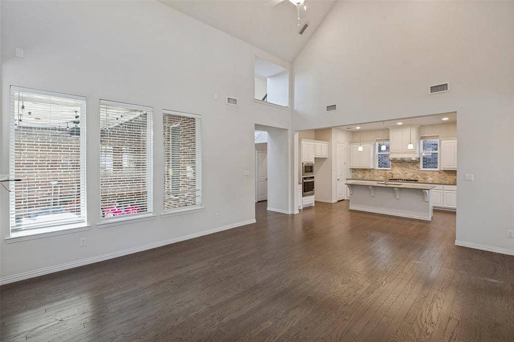 Expansive living area with vaulted ceilings, wood-finish flooring, three oversized windows, and an island kitchen featuring white cabinetry and built-in appliances