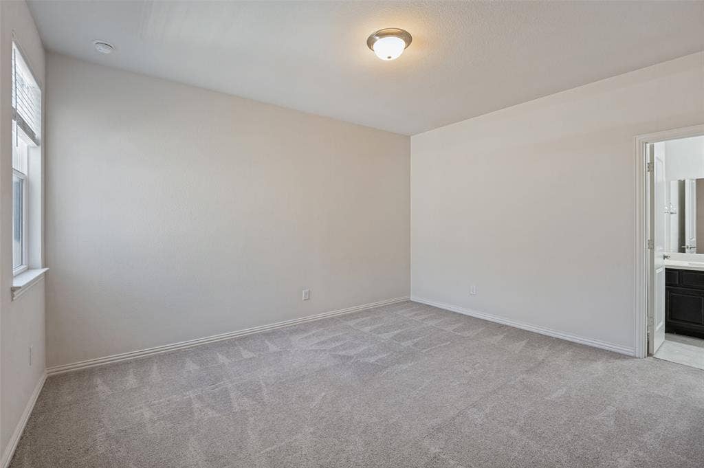 Neutral-toned interior space featuring wall-to-wall carpeting, single-pane window with sill, flush-mount ceiling lighting, and a doorway revealing a vanity with dark cabinetry