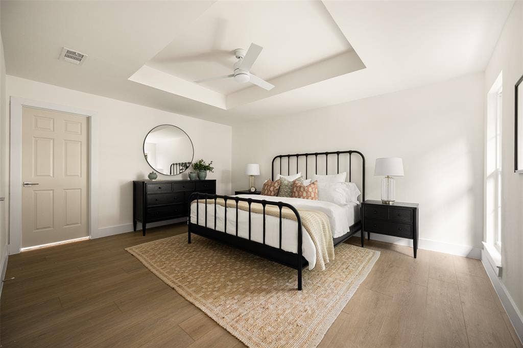 Beautifully updated primary bedroom featuring a tray ceiling, modern ceiling fan, warm wood-like flooring, and abundant natural light that creates a bright, inviting atmosphere