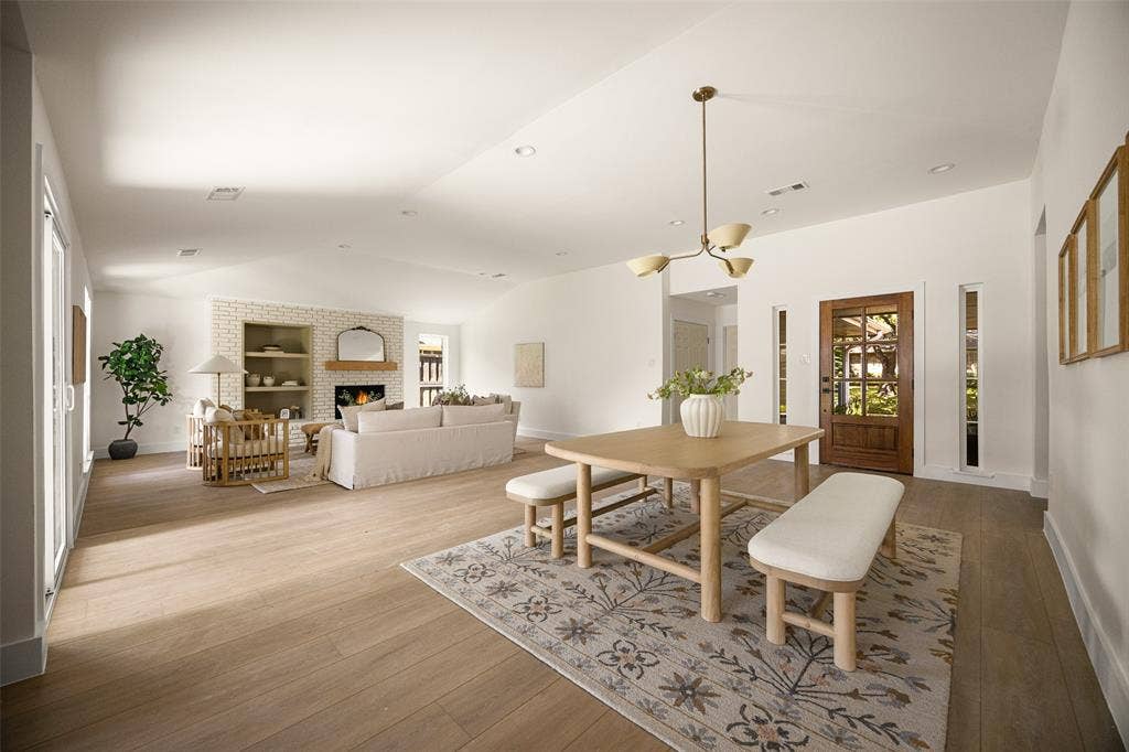 Open living and dining areas with vaulted ceilings, wood-like flooring, modern lighting, and a brick fireplace with built-in shelving.