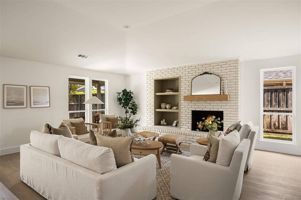 Bright and inviting living area featuring vaulted ceilings, a beautifully updated brick fireplace with wood mantel, built-in shelving, and large windows overlooking the backyard.