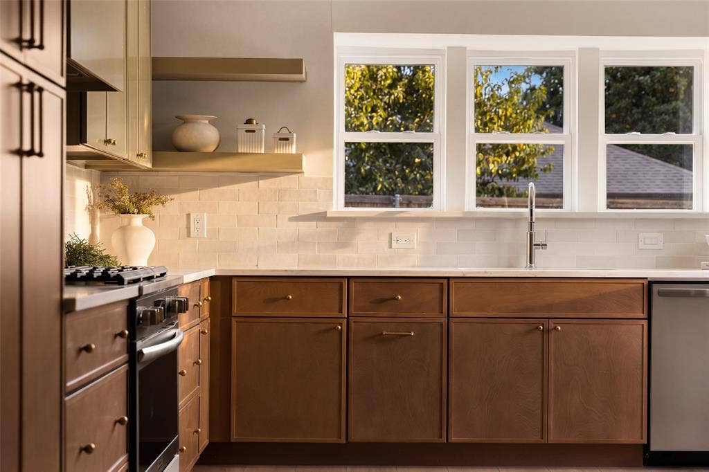 Sophisticated, designer-inspired kitchen featuring rich wood cabinetry, elegant stone-look countertops, artisan-style tile backsplash, warm brass accents, stainless steel appliances, and abundant natural light.