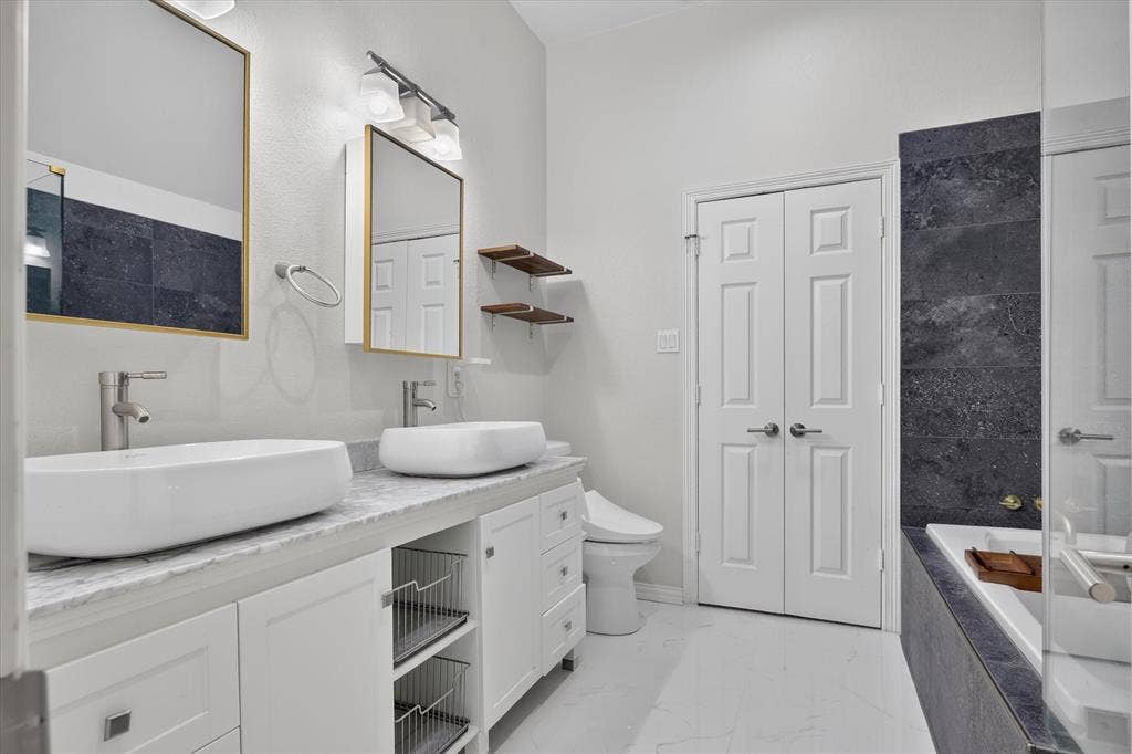 1825 Bachman Court, Plano, TX 75075 Dual vanity with rectangular vessel sinks, stone-finish countertop, white cabinetry with built-in wire storage, wall-mounted light fixtures, and a soaking tub featuring dark-toned stone-finish tile surround