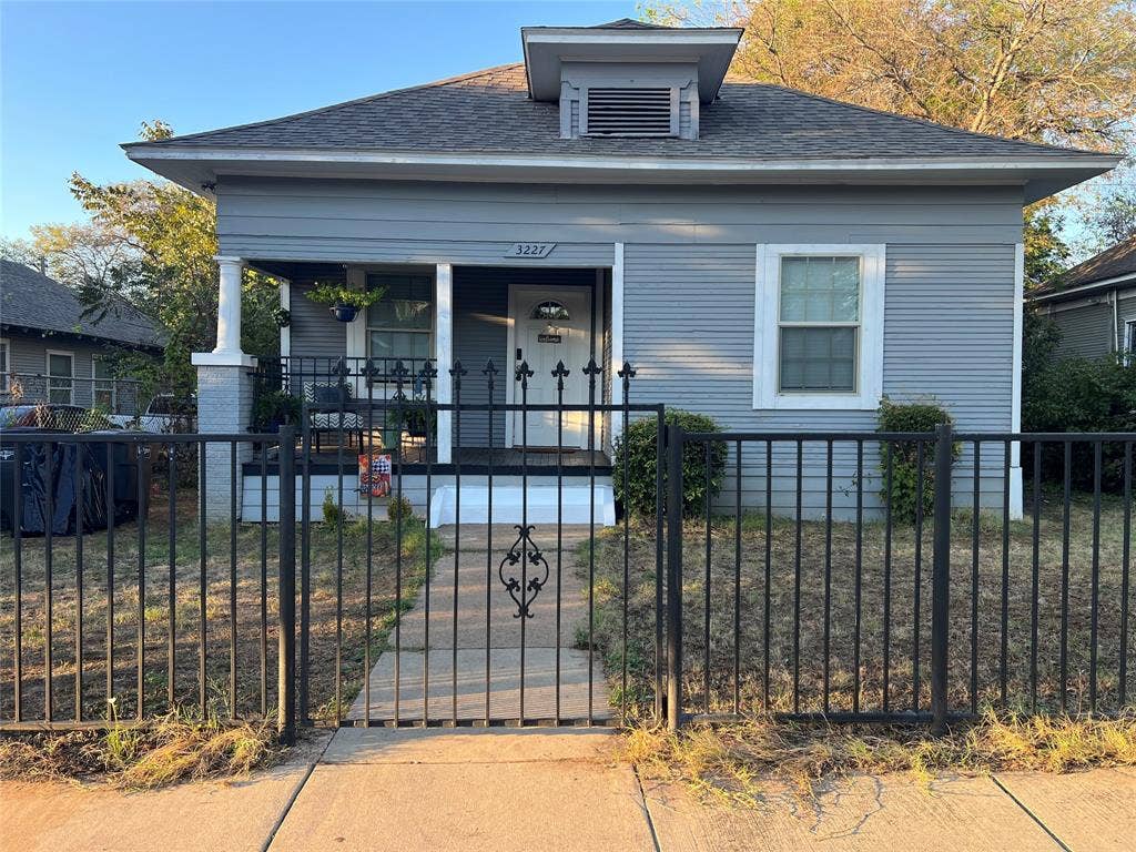 3227 Avenue I, Fort Worth, TX 76105 Exterior featuring light blue siding, a covered porch with white columns, a dormer with a vent, and a black wrought iron fence with an ornamental gate