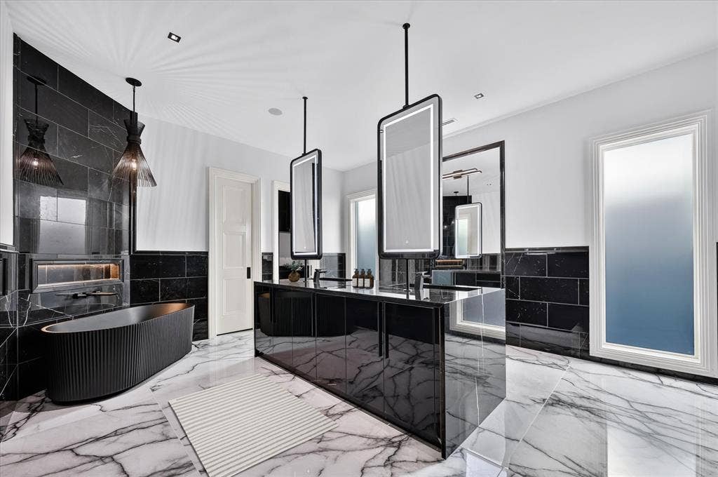 6404 Lakehurst Avenue, Dallas, TX 75230 Freestanding soaking tub, floor-to-ceiling black stone-finish wall, dual vanity with integrated sinks, illuminated mirrors, and white marble-look flooring