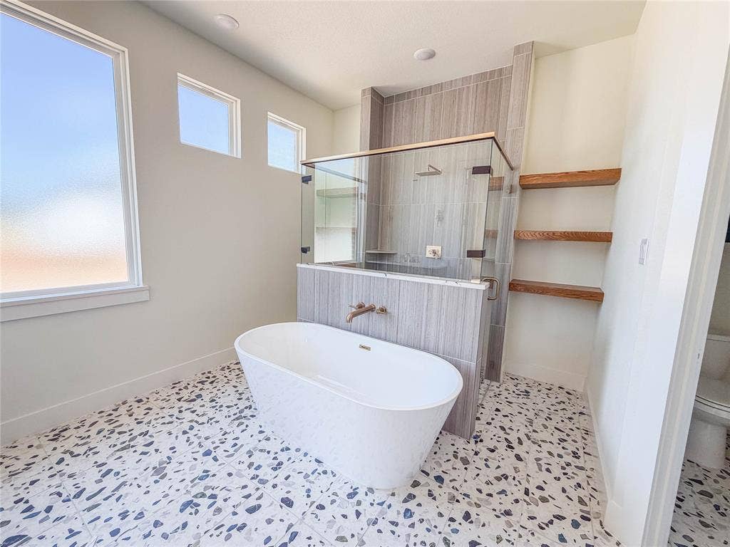 3860 Jacks Loop, Midlothian, TX 76065 Freestanding soaking tub with a wall-mounted bronze faucet, glass-enclosed shower with tile surround, wood-finish shelving, and patterned tile flooring