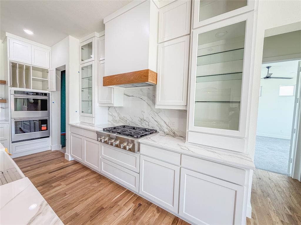 3860 Jacks Loop, Midlothian, TX 76065 White cabinetry with glass-front display cabinets, light wood-finish flooring, stainless steel gas range with a matching range hood featuring a wood accent, and integrated stainless steel double oven