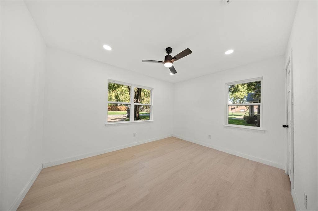 Neutral-toned interior space featuring light wood-finish flooring, recessed lighting, dual windows, and a matte black ceiling fan