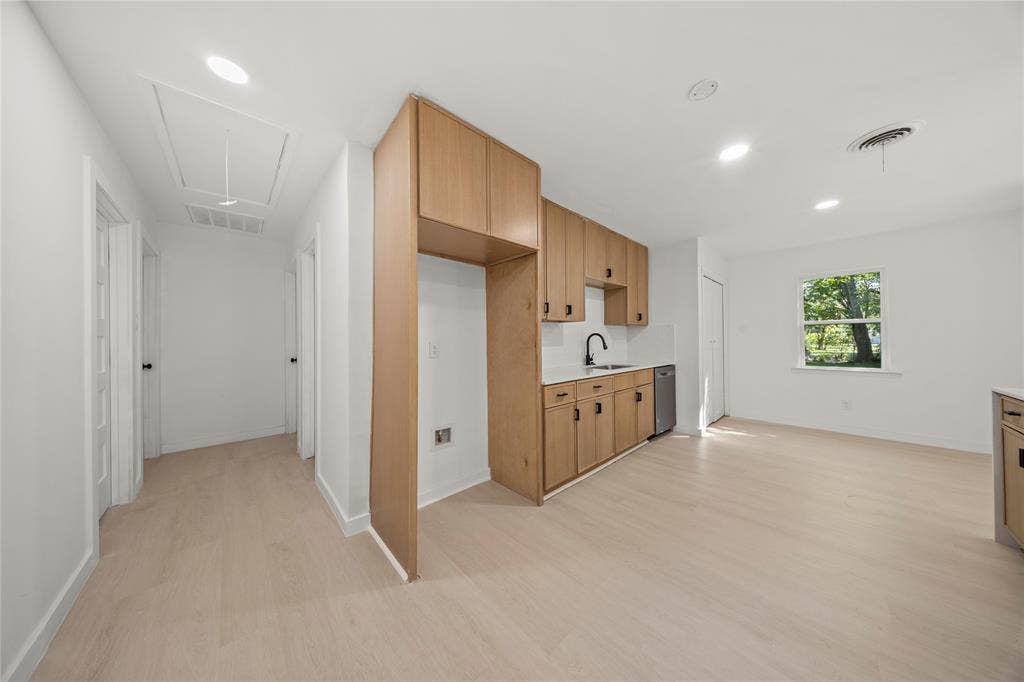 Kitchen featuring light wood-finish cabinetry, neutral solid-surface countertops, a single-basin sink with a matte black faucet, integrated dishwasher, and light-toned wood-finish flooring