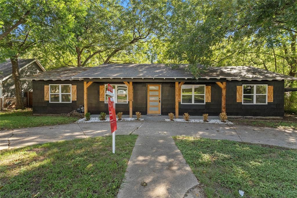Single-story exterior featuring dark siding, decorative wood-finish shutters, a wood-finish front door with horizontal glass panels, and a covered front porch with timber-frame support posts