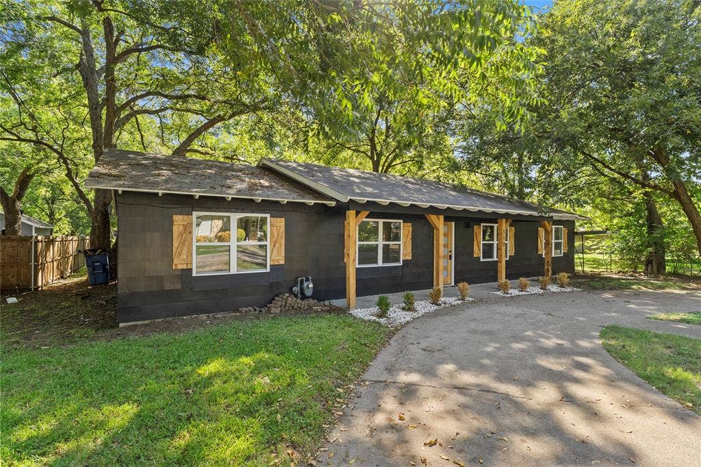 Single-story exterior featuring dark siding, decorative wood shutters, a covered front porch with timber support posts, and a paved driveway