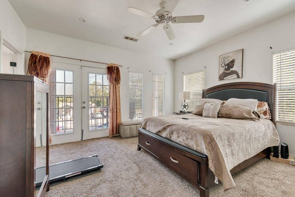 Spacious primary bedroom with French doors opening to a private balcony, ceiling fan, plantation shutters, wood-finish flooring, and carpet accents