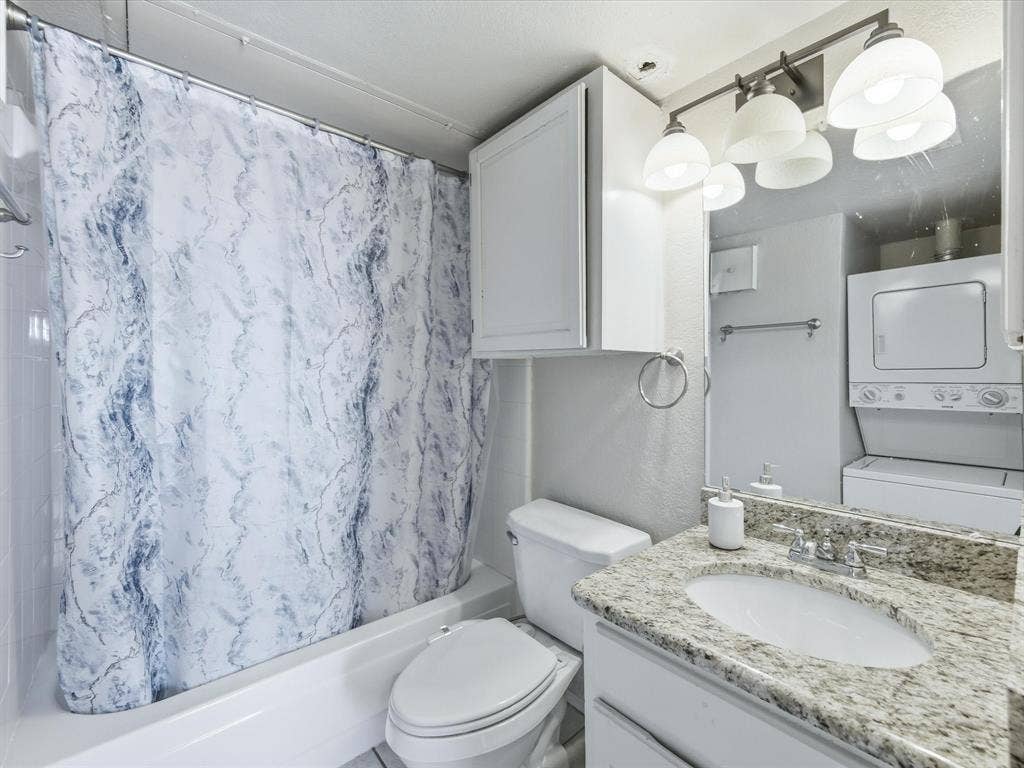 3129 Sondra Drive, #206, Fort Worth, TX 76107 Bathroom featuring a shower-tub combination, a toilet, a vanity with a granite countertop, and a stacked washer and dryer