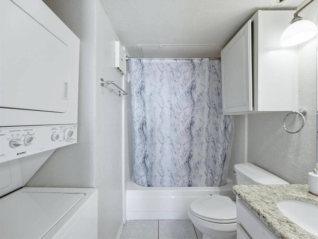 3129 Sondra Drive, #206, Fort Worth, TX 76107 Bathroom featuring a white stacked washer and dryer, a tub-shower combination with a patterned curtain, a toilet, and a vanity with a light-colored granite countertop and integrated sink