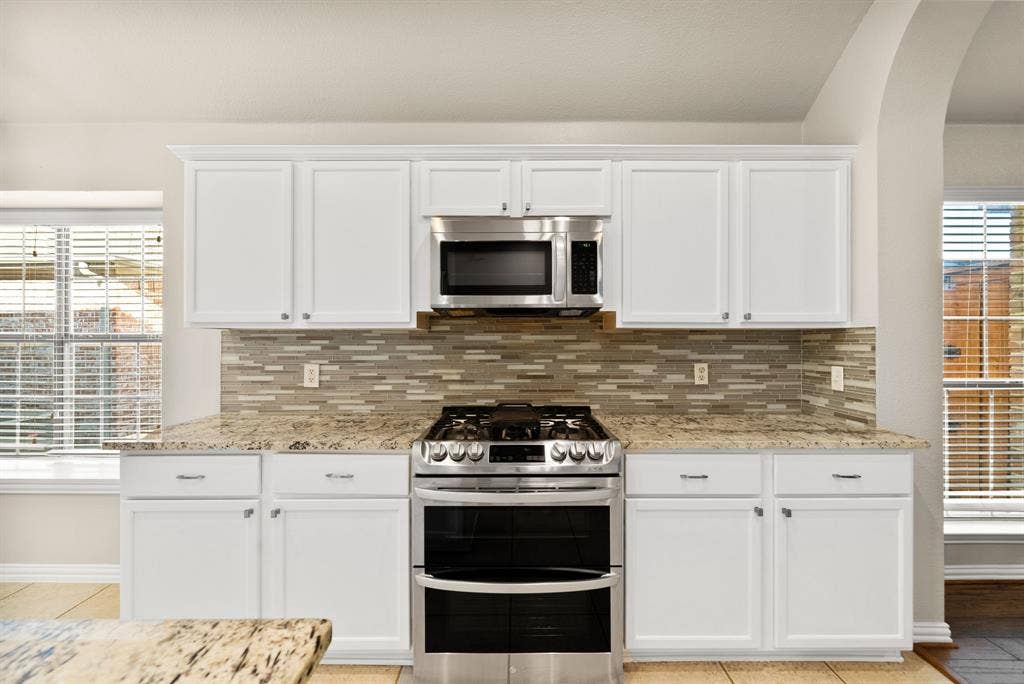 242 Cox Drive, Fate, TX 75087 White cabinetry, stainless steel oven with gas range, stainless steel microwave, stone countertops, and a tiled backsplash