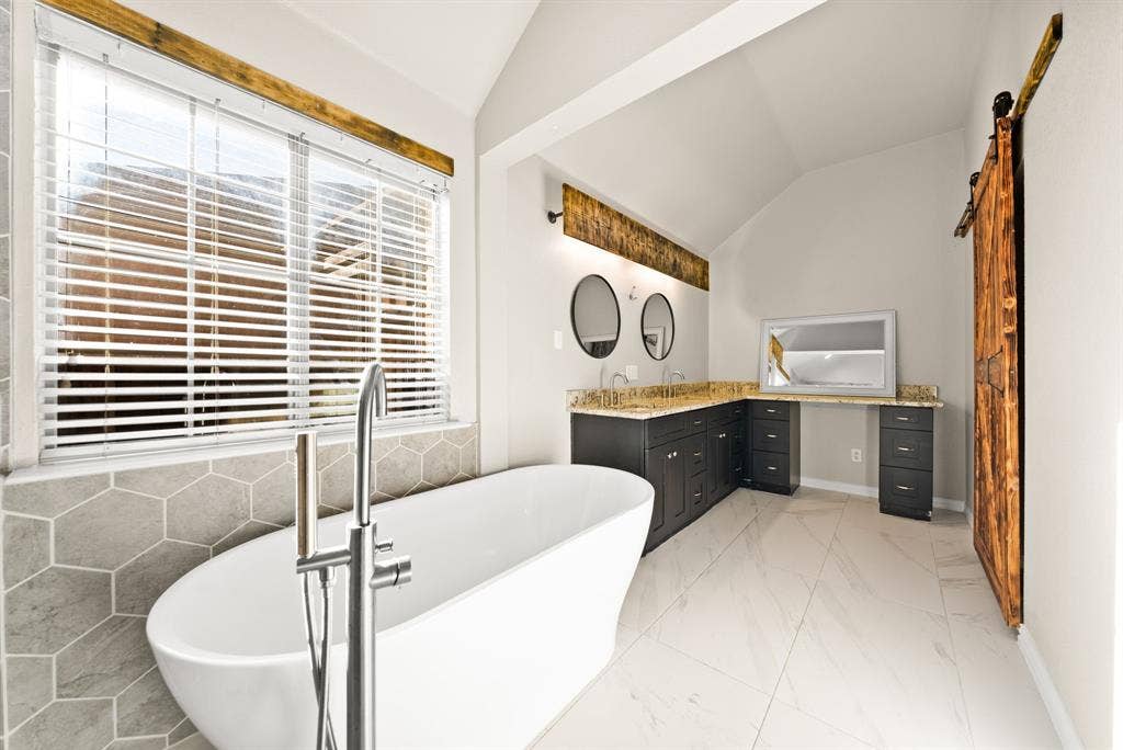 242 Cox Drive, Fate, TX 75087 Spa-inspired primary bathroom featuring a freestanding soaking tub with floor-mounted filler, dual vanity with black cabinetry and wood-finish countertops, vessel sinks, polished nickel hardware, and large-format tile flooring