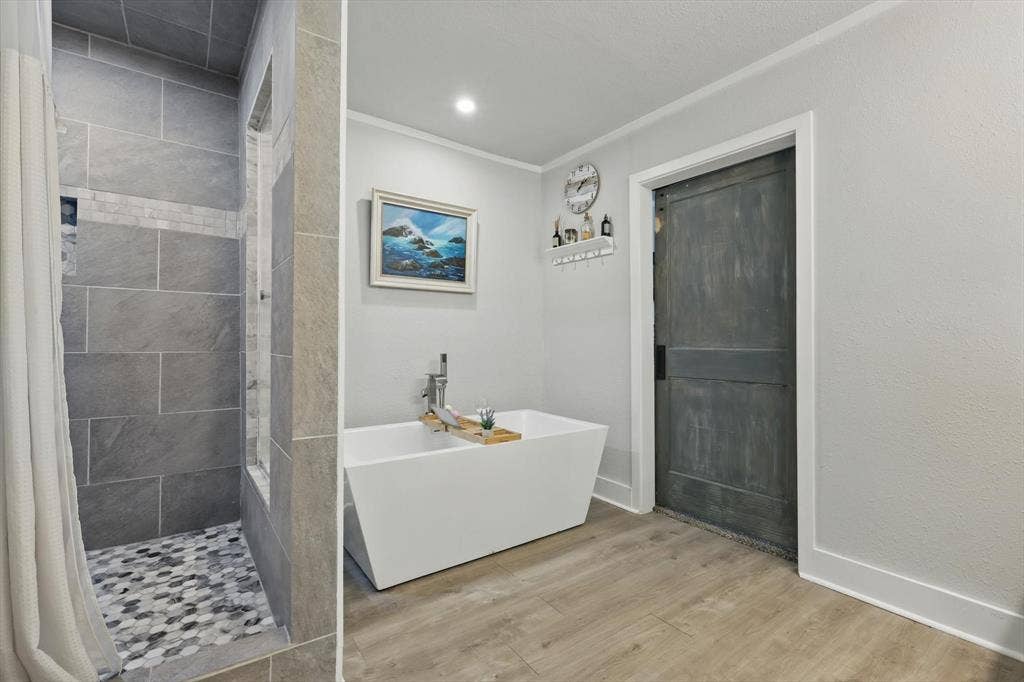 5247 Fm 1569, Farmersville, TX 75442 Spa-inspired primary bathroom featuring a freestanding soaking tub, walk-in shower with large-format tile surround and mosaic floor detail, wood-finish flooring, recessed lighting, and soft gray wall finishes complemented by coastal artwork