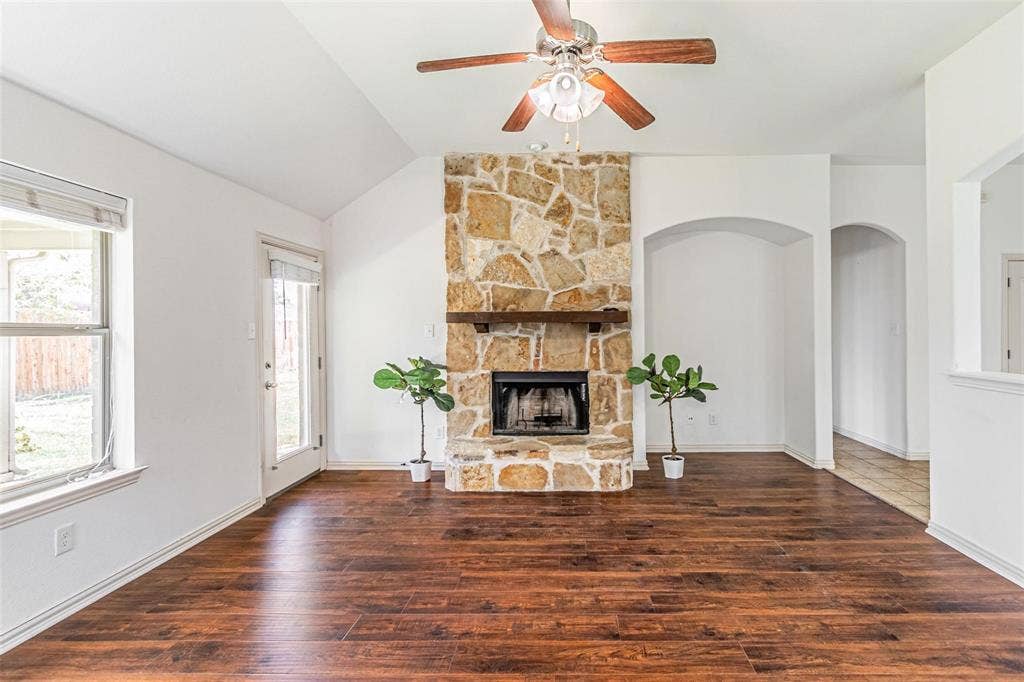 Vaulted ceilings, wood-finish flooring, stone fireplace with hearth and wood mantel, ceiling fan with integrated lighting, and an arched entryway