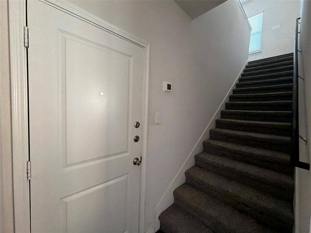 Entryway featuring a solid panel door with metallic hardware, neutral wall paint, and a carpeted staircase with a metal handrail