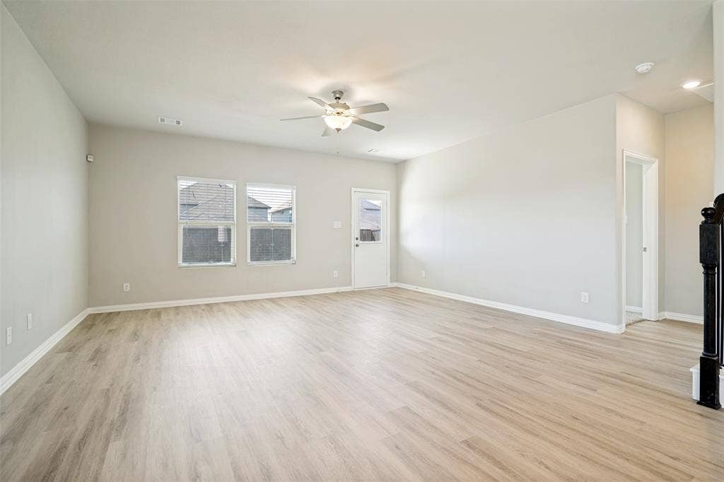 Spacious room featuring light-toned wood-finish flooring, multiple windows, a ceiling fan with integrated lighting, and a white door with an exterior light