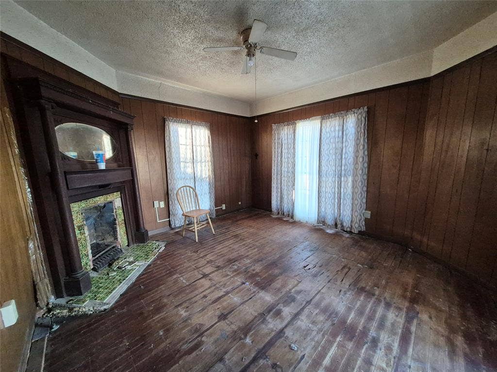 Wood paneling walls, wood-finish flooring, two window openings, ceiling fan, and a fireplace with a wood mantel
