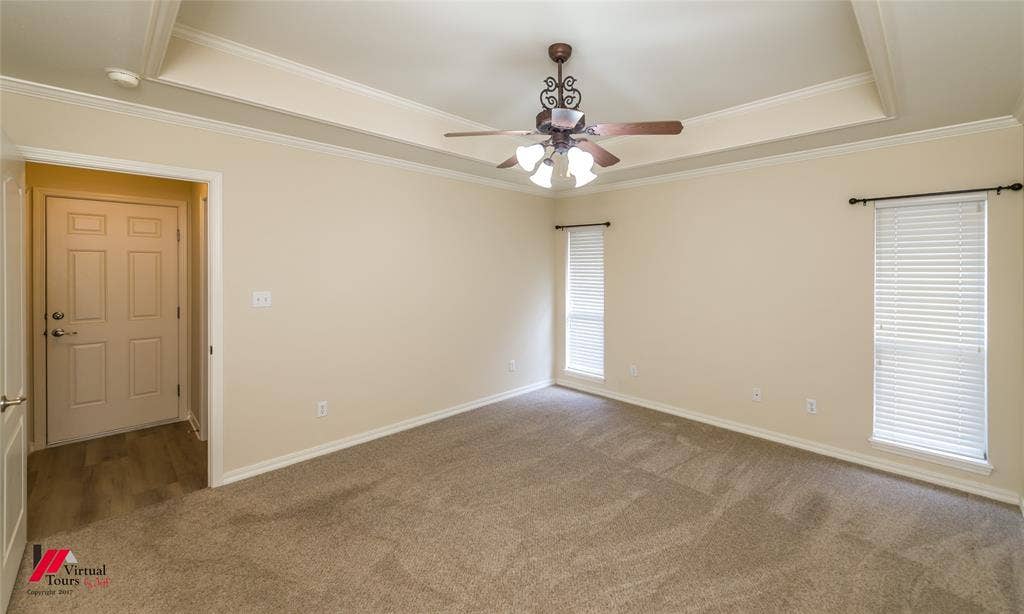 Tray ceiling featuring a decorative central ceiling fan with multi-light fixture, neutral carpeting, and dual windows with fitted blinds
