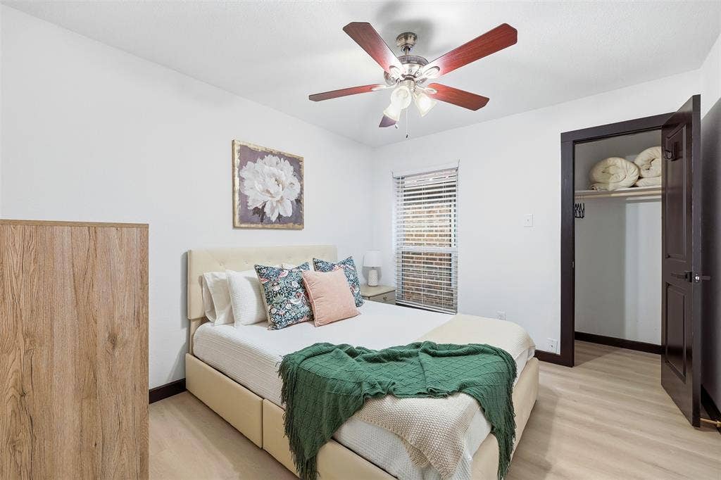 Light wood-finish flooring, ceiling fan with integrated lighting, window with blinds, and built-in closet with shelving and hanging rod