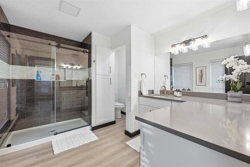 Spacious bathroom featuring a glass-enclosed shower with dark tile surround, a dual vanity with light countertops, and wood-finish flooring