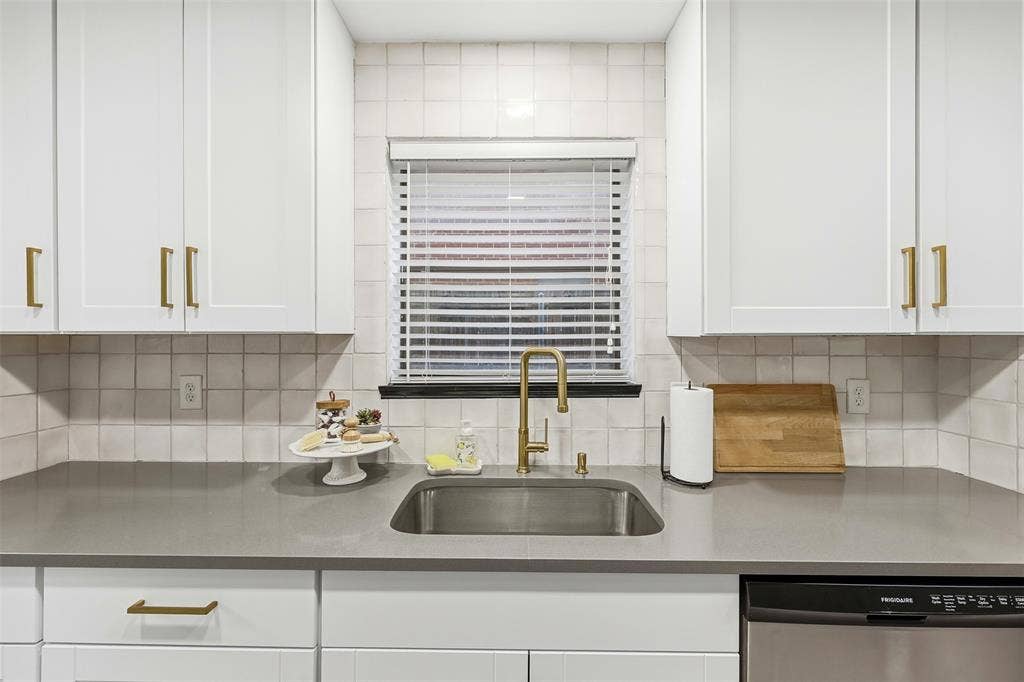 White shaker-style cabinetry with gold-finish hardware, light-toned square tile backsplash, stainless steel sink with gold-finish faucet, and a stainless steel Frigidaire dishwasher