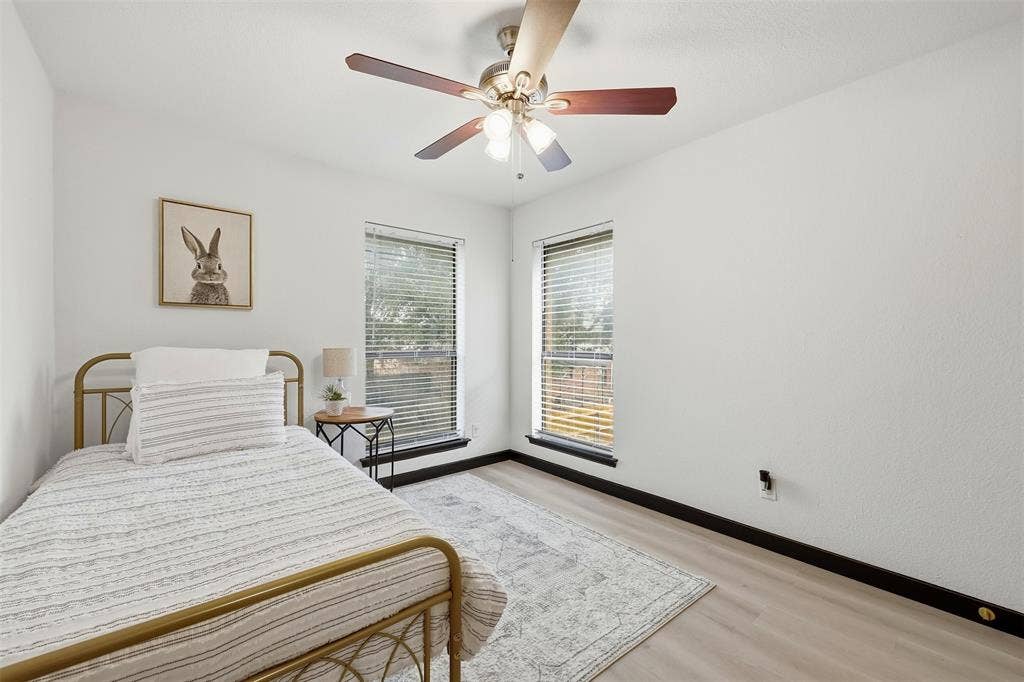 Light-toned wood-finish flooring, two large windows with blinds, ceiling fan with integrated lighting, and white painted walls