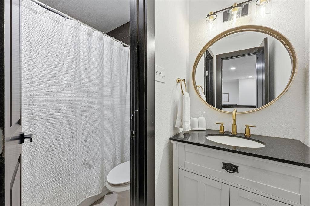 Vanity with a black countertop, gold-finish fixtures, and an oval mirror, complemented by an adjacent shower