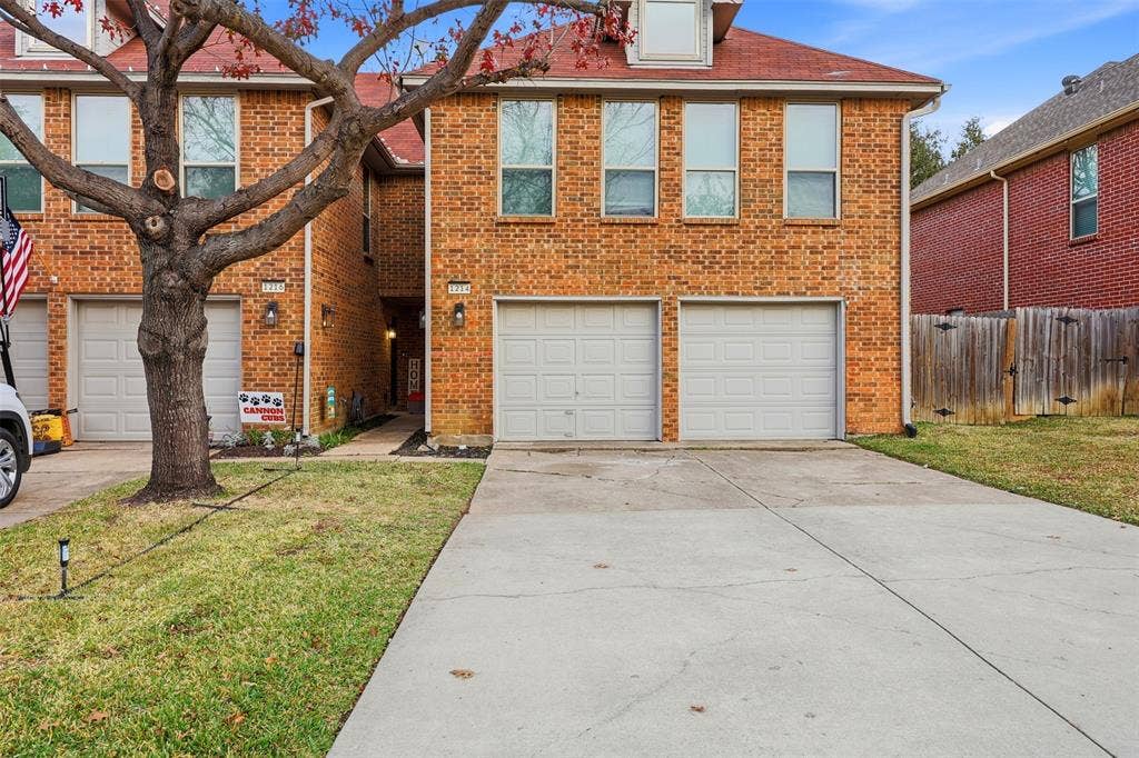 Brick exterior, dual single-car garages, red shingle roof, concrete driveway, and established front yard tree