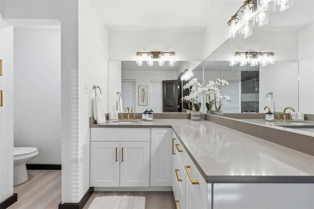 Dual vanity with undermount sinks, expansive countertop, white cabinetry with gold-tone hardware, and mirrored wall
