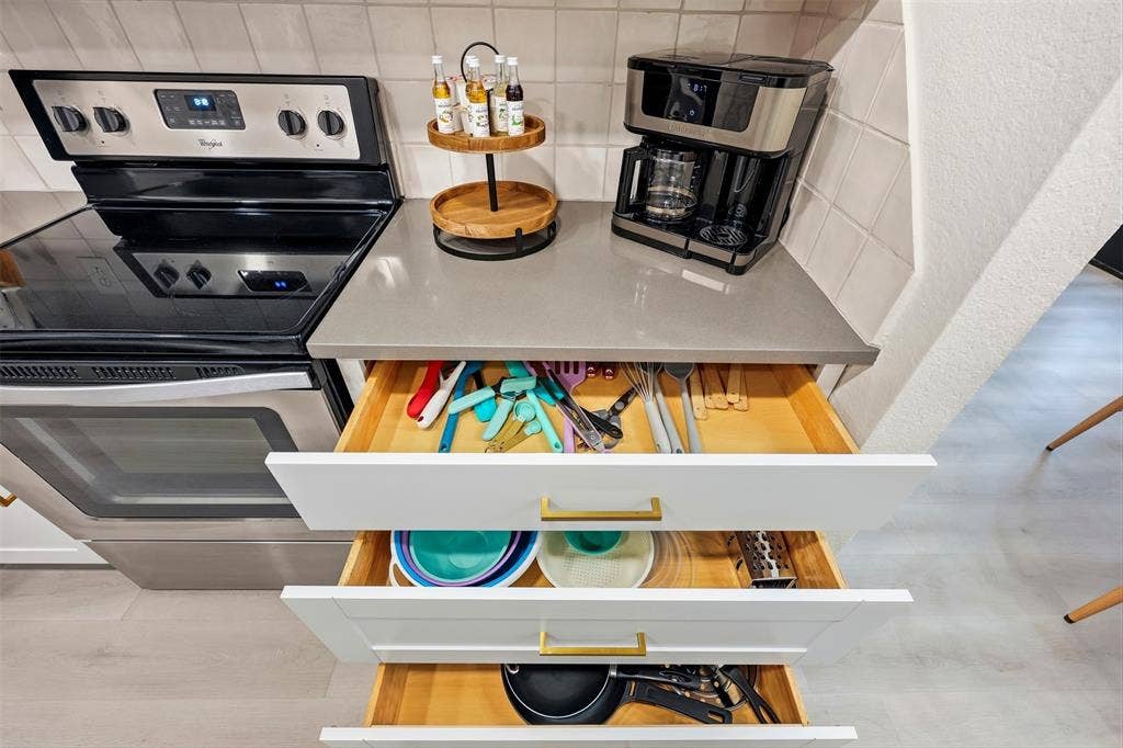 Stainless steel range, light-toned countertop, white cabinetry with gold-tone hardware, white tile backsplash, light wood-finish flooring