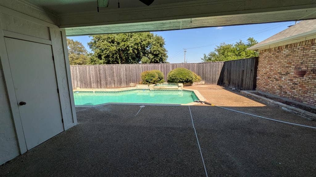 Outdoor covered patio with exposed aggregate surface, built-in swimming pool, surrounding wood fencing, and brick exterior wall