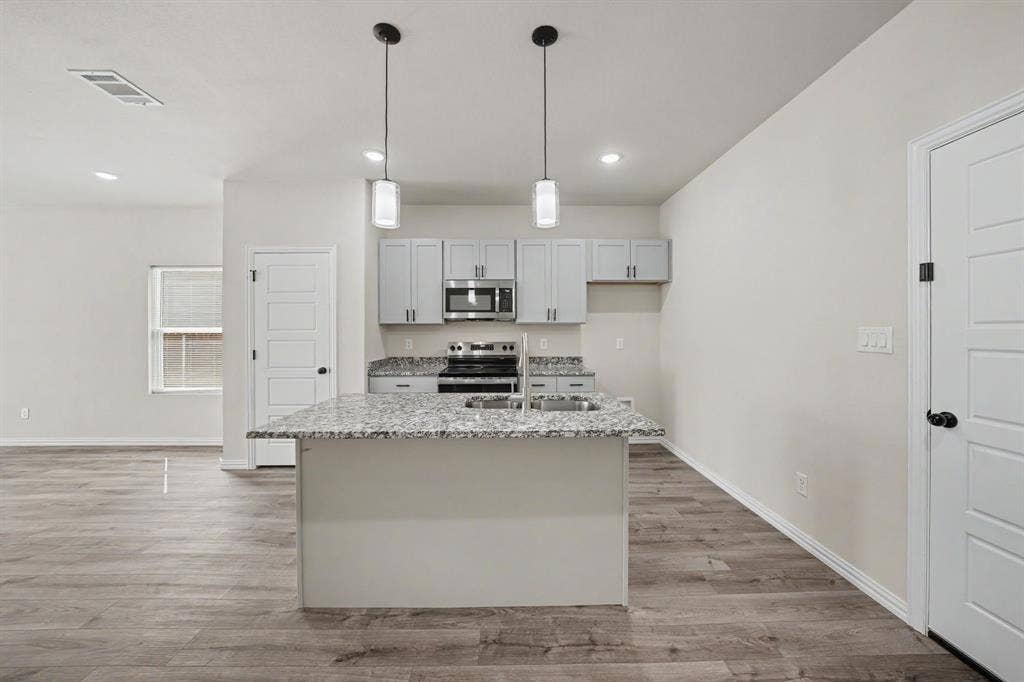 Kitchen featuring an island with a built-in sink, pendant lighting, light gray cabinetry, stainless steel appliances, and wood-finish flooring