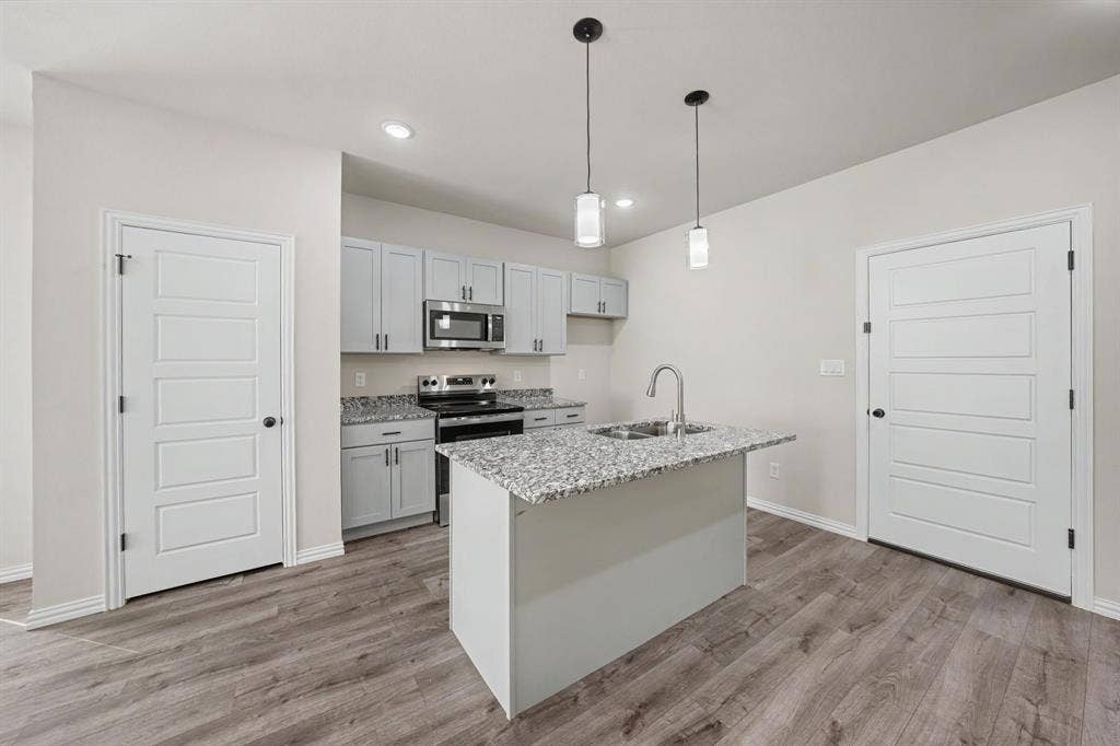 Kitchen featuring wood-finish flooring, light gray cabinetry, stainless steel appliances, a center island with a sink, and pendant lighting