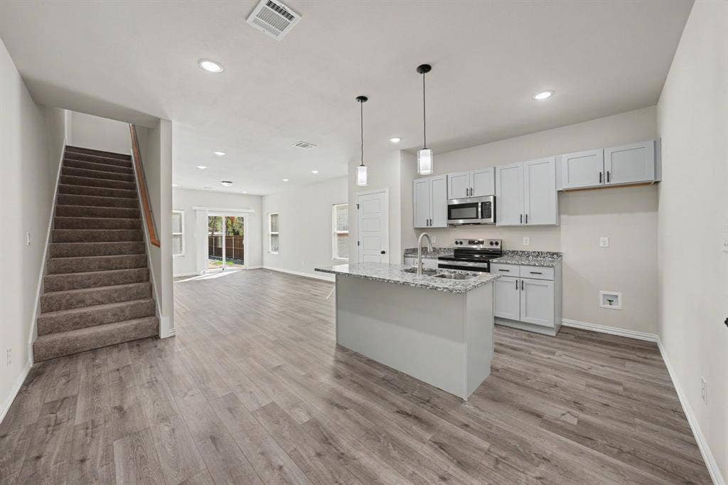 Open-concept design featuring wood-finish flooring, a kitchen island with pendant lighting, light gray cabinetry, stainless steel appliances, and a carpeted staircase