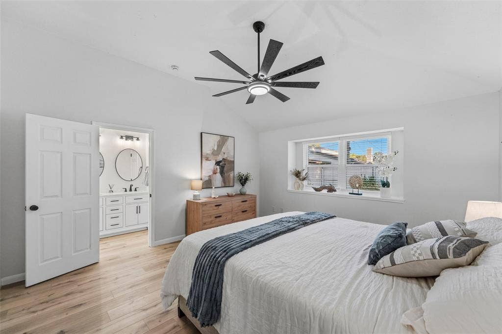 Vaulted ceiling with a contemporary ceiling fan, wood-finish flooring, a rectangular window with horizontal blinds, and an attached bath with a double vanity