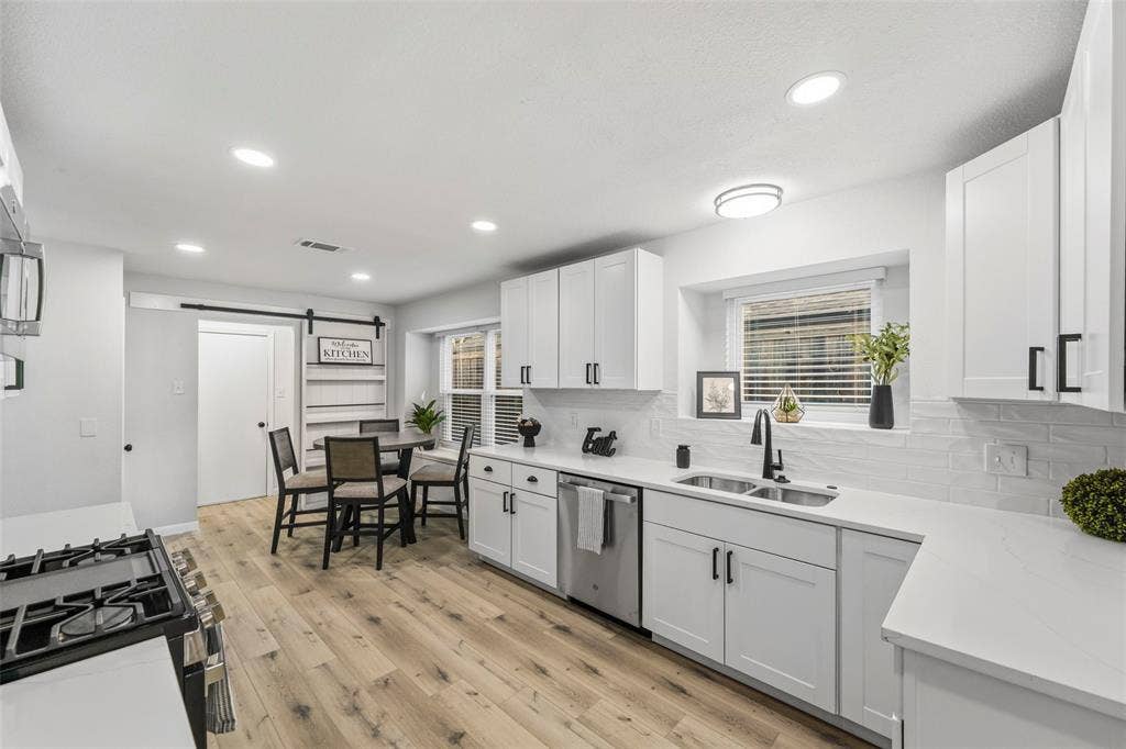White cabinetry with matte black hardware, dual basin stainless steel sink, light wood-finish flooring, recessed lighting, and a barn door entry