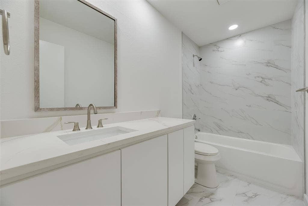 1903 Clemson Drive, Richardson, TX 75081 White vanity with integrated sink, brushed nickel faucet, large framed mirror, white bathtub with wall-mounted shower, and marble-look tile surround