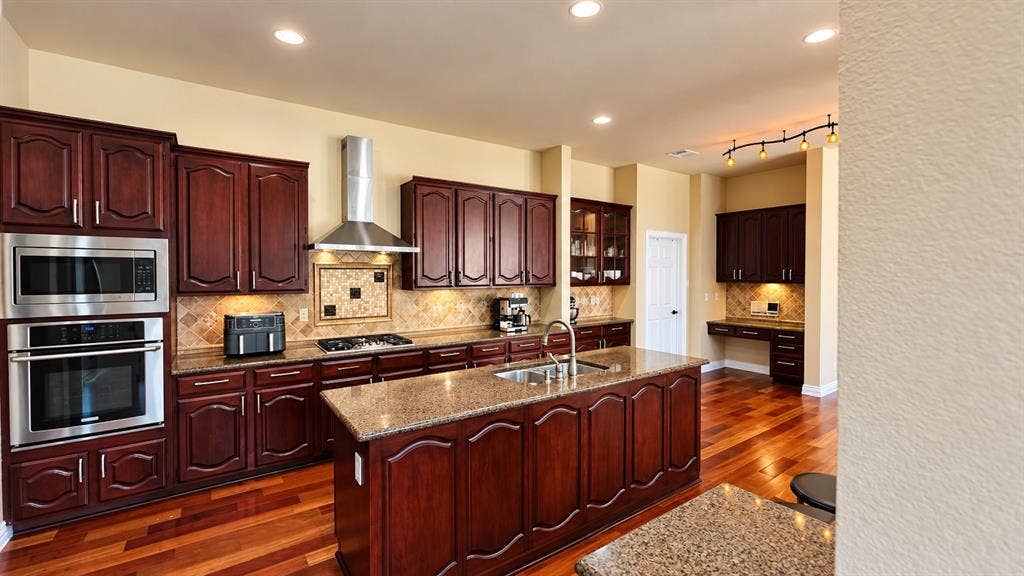 4505 Southgate Drive, Plano, TX 75024 Kitchen featuring wood-finish flooring, dark wood cabinetry, stainless steel appliances, a center island with a sink, and recessed lighting