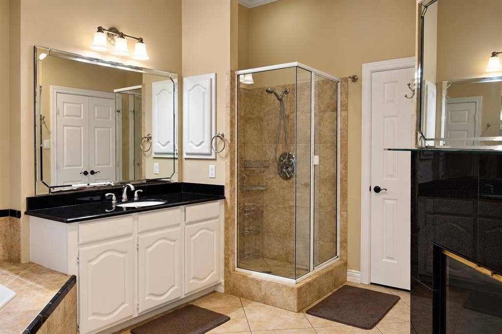 4505 Southgate Drive, Plano, TX 75024 Tile flooring, white vanity with black countertop, glass-enclosed shower, wall-mounted vanity lights, and a large wall mirror