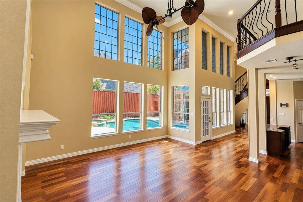 4505 Southgate Drive, Plano, TX 75024 Two-story living area with high ceilings, featuring wood-finish flooring, an ornate staircase, and multiple windows providing ambient light and a pool setting