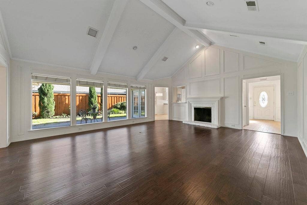 16353 Lauder Lane, Dallas, TX 75248 Dark wood-finish flooring, vaulted ceiling with exposed beams, white paneling accents, classic white fireplace, and a wall of windows overlooking a private yard