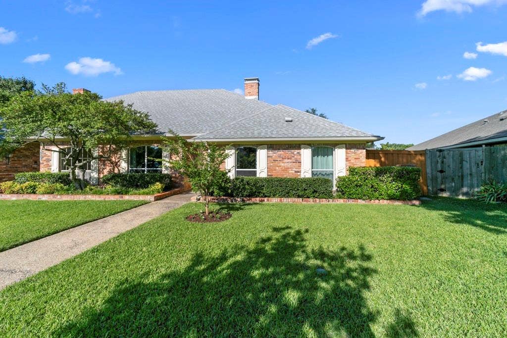 16353 Lauder Lane, Dallas, TX 75248 Brick exterior with a gray shingled roof, featuring white window shutters, mature landscaping, and a concrete pathway