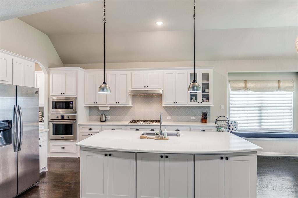 1481 Ramsbury Lane, Prosper, TX 75078 White shaker cabinetry, stainless steel appliances, central island with solid surface countertop, white subway tile backsplash, and vaulted ceiling