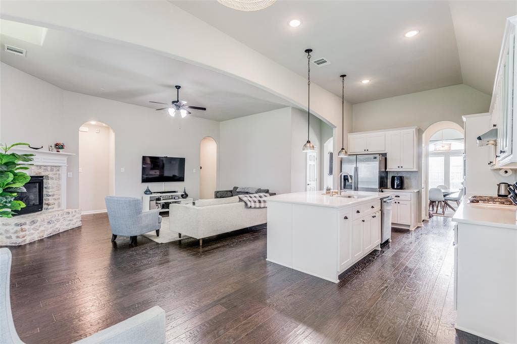 1481 Ramsbury Lane, Prosper, TX 75078 Open-concept living space featuring wood-finish flooring, a white kitchen island with sink, stainless steel appliances, a stone-clad fireplace, and arched doorways